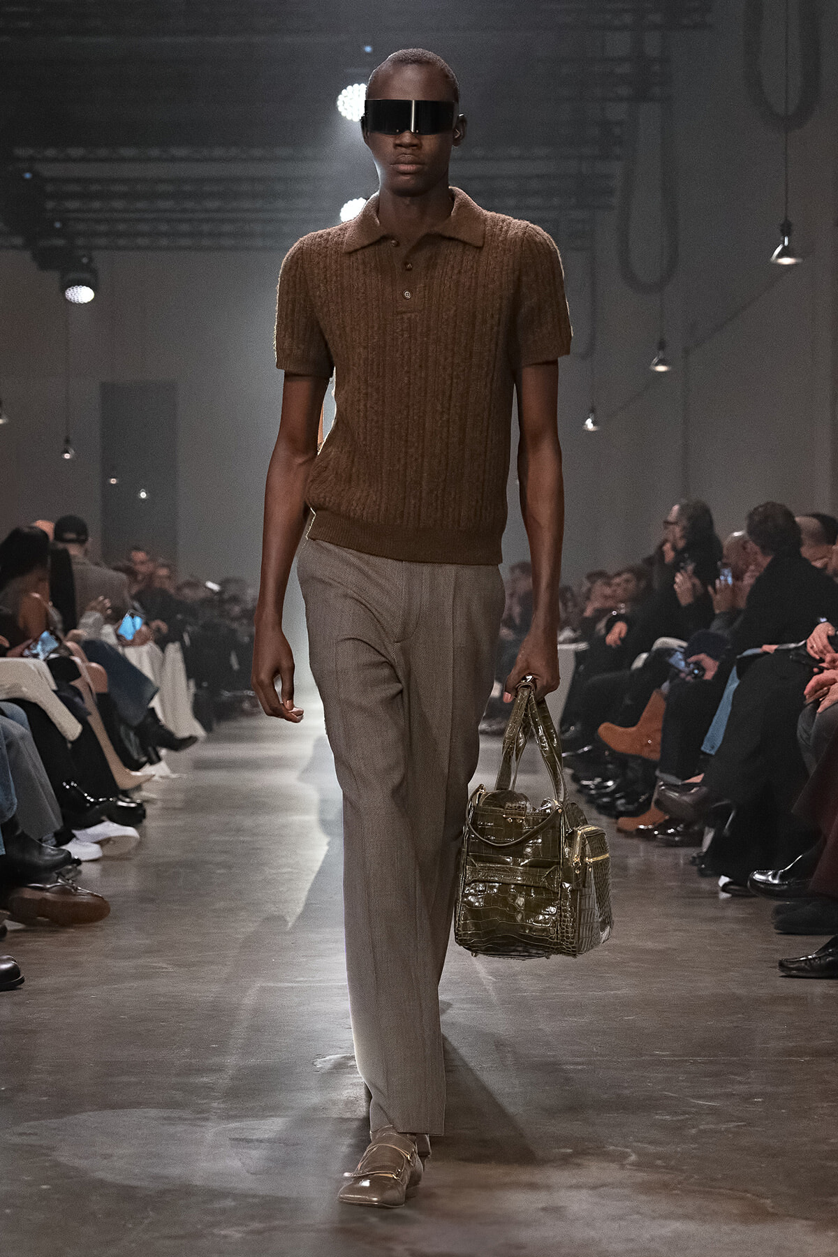 Male model on runway wearing brown knit polo, beige trousers, oversized black visor sunglasses, and carrying a metallic handbag; audience seated along the catwalk.