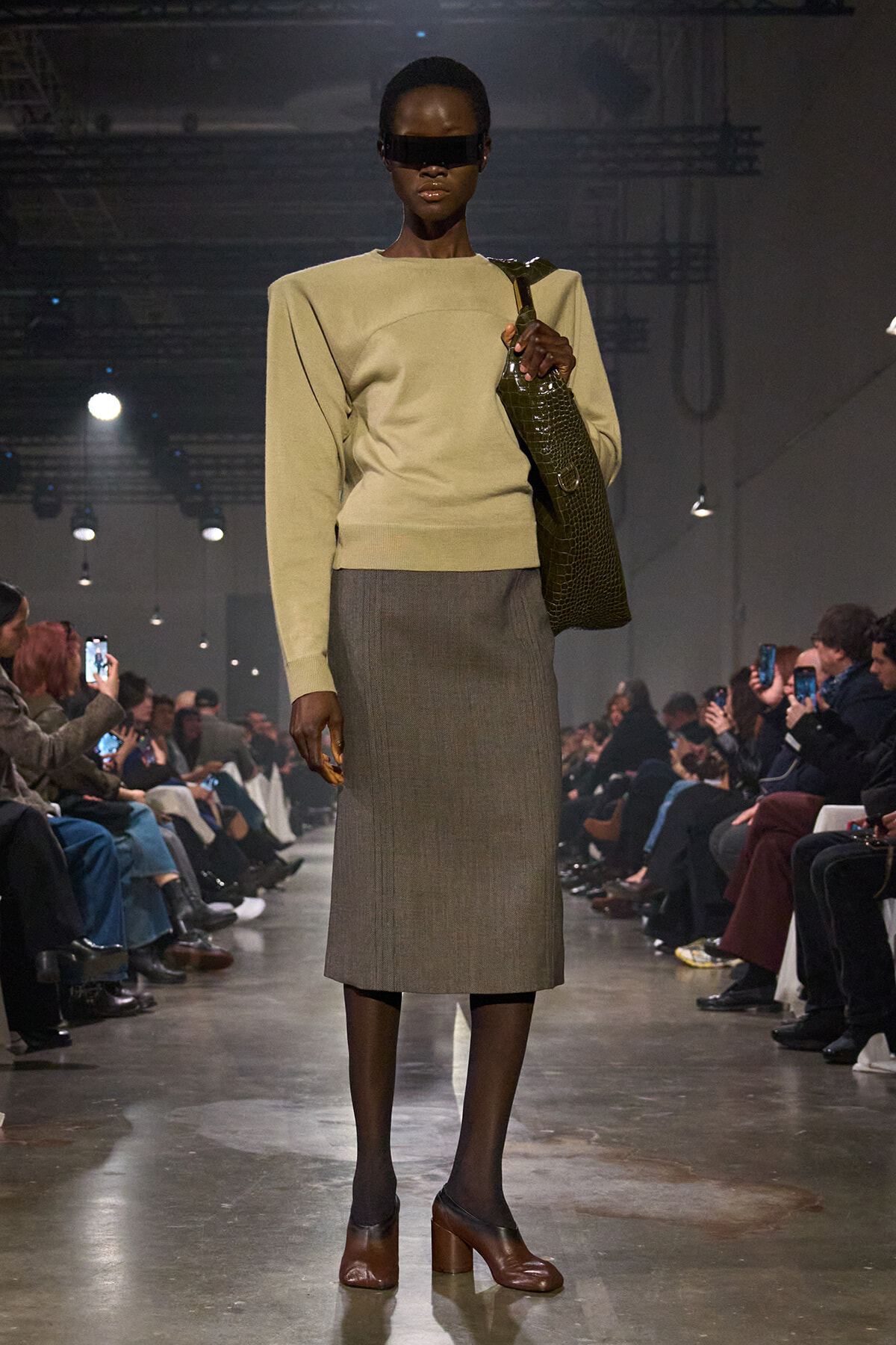 Black model on a runway wearing a beige sweater, gray midi skirt, brown heels, and a dark green crocodile bag; audience filming in the background.