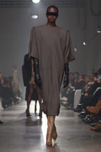 Model walking a runway in a loose taupe dress with oversized sleeves, black gloves, and tan knee-high boots under studio lights.”],