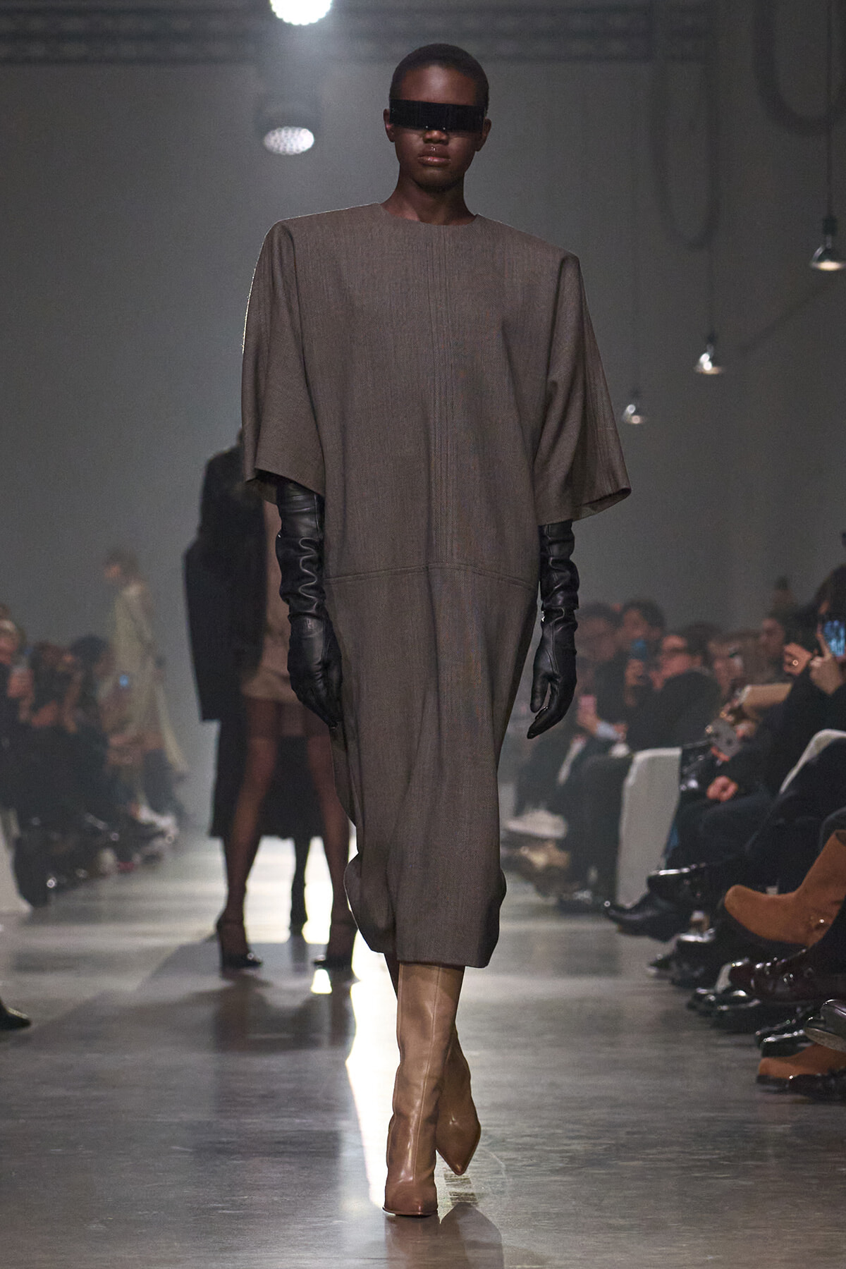 Model walking a runway in a loose taupe dress with oversized sleeves, black gloves, and tan knee-high boots under studio lights.”],