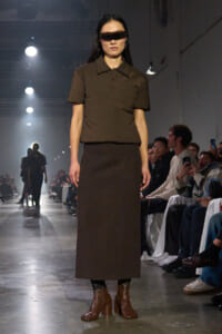 Runway model wearing a brown polo shirt, long brown skirt, and brown heeled boots, with a dark rectangular visor over her eyes.