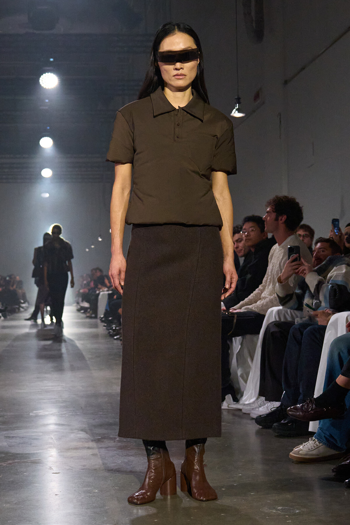 Runway model wearing a brown polo shirt, long brown skirt, and brown heeled boots, with a dark rectangular visor over her eyes.