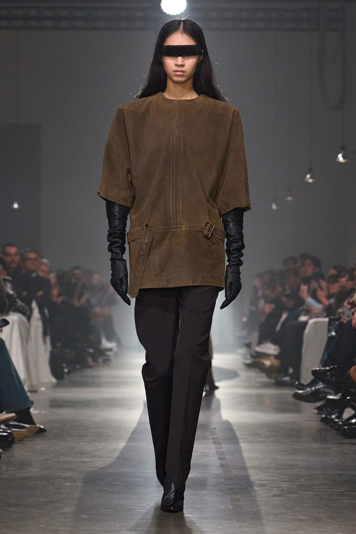 Model walks the runway wearing a brown suede tunic with wide sleeves, belted at the waist, paired with black pants and long black gloves; eyes covered with a black blindfold-like band for the show theme.