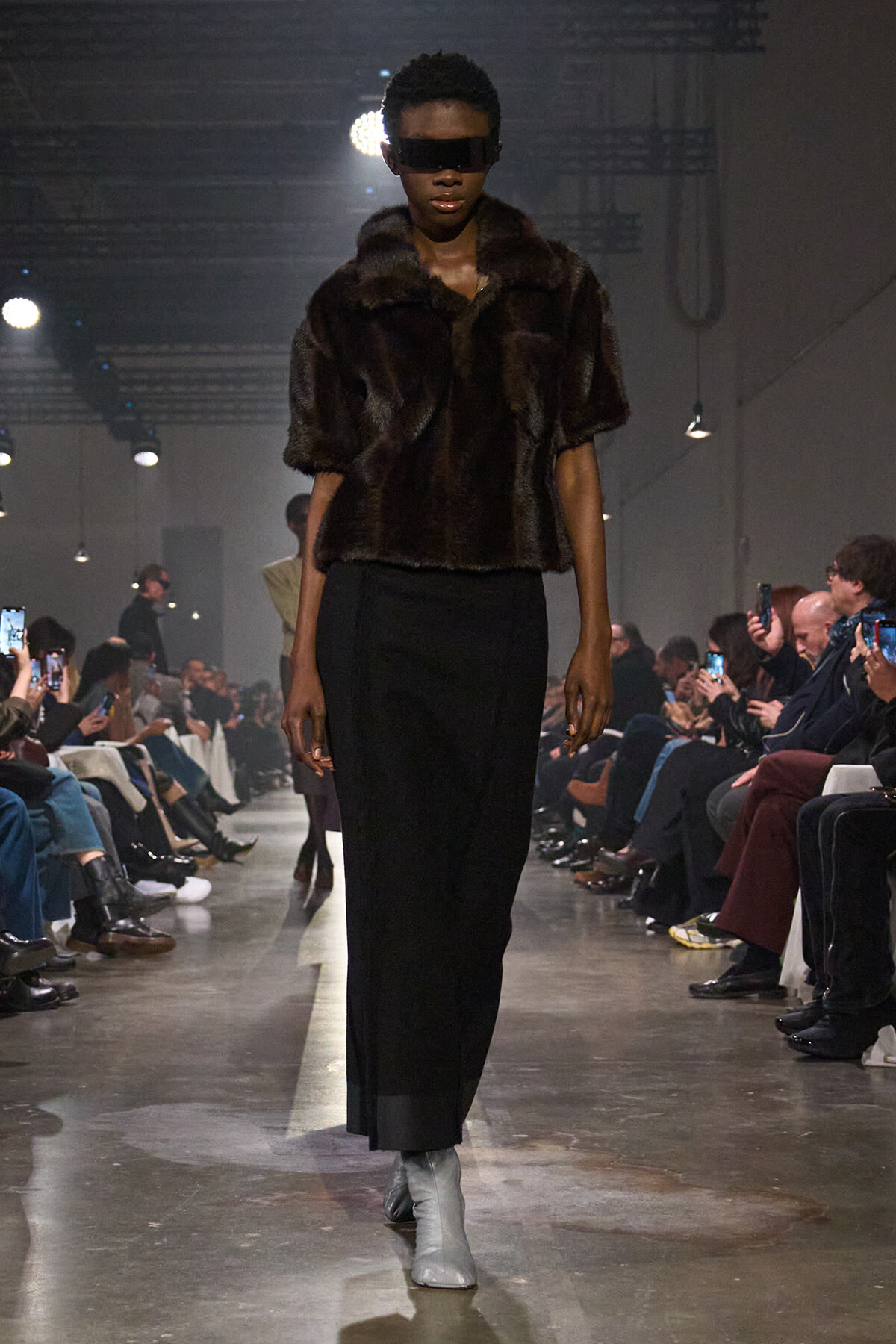 Model walks the runway in a dark fur cropped coat, black wide-leg pants, and light gray ankle boots; audience rows line the seated sides.