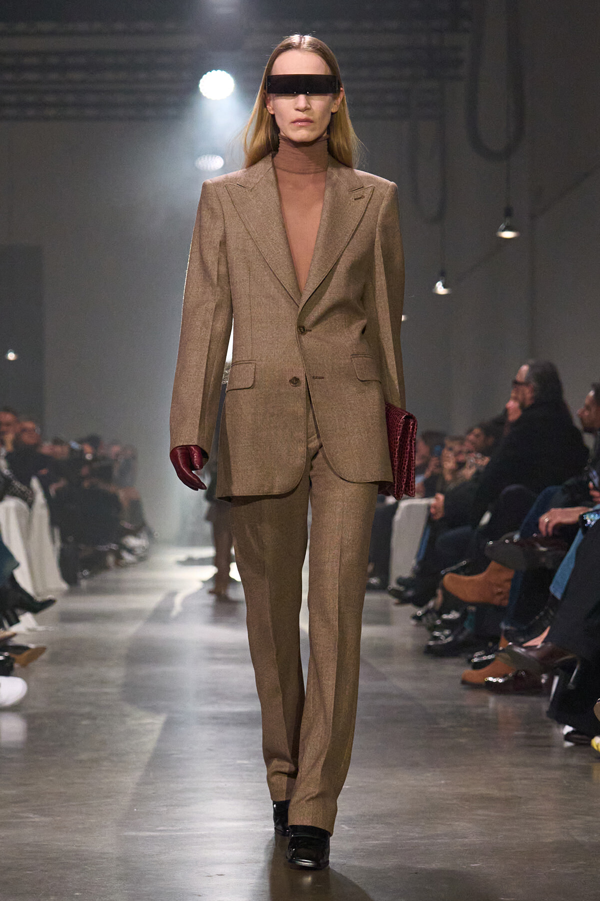 Model walks runway in a tan oversized suit with a brown turtleneck and futuristic black visor sunglasses, red gloves, and a red clutch.