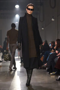 Model on the runway wearing a long black coat over a brown turtleneck, black gloves, knee-high boots, and a futuristic visor eyewear behind dark sunglasses shows a sci‑fi look.