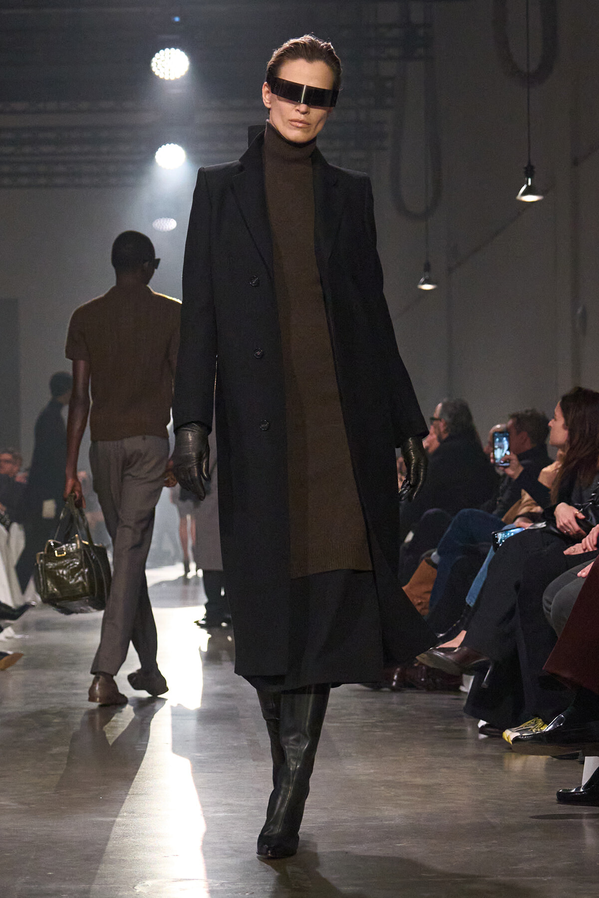 Model on the runway wearing a long black coat over a brown turtleneck, black gloves, knee-high boots, and a futuristic visor eyewear behind dark sunglasses shows a sci‑fi look.