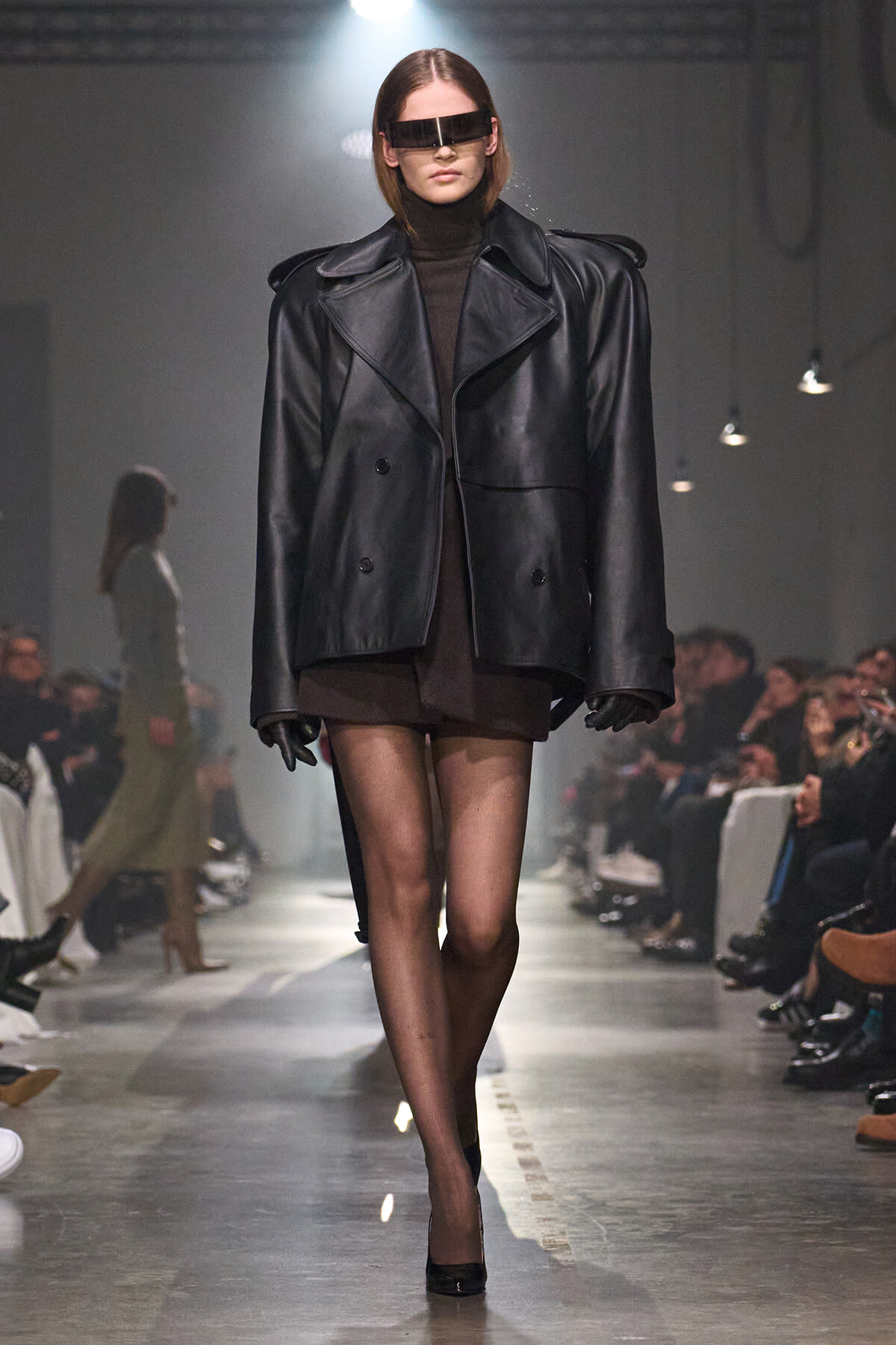 Model on a runway wearing an oversized black leather blazer over a brown dress, sheer tights, and futuristic visor sunglasses.