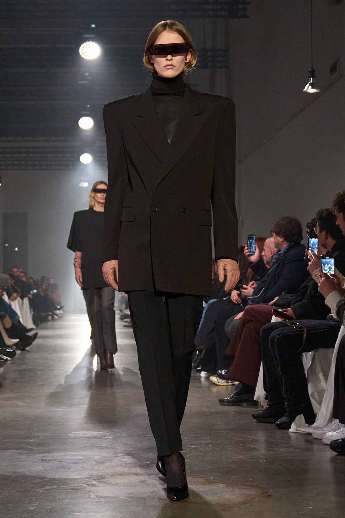 Model on a runway wearing an oversized black blazer and matching pants with a black turtleneck and futuristic rectangular visor sunglasses.