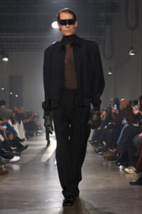 Male model on a runway in an all-black ensemble with a sheer top, zip jacket, oversized visor sunglasses, and gloves.