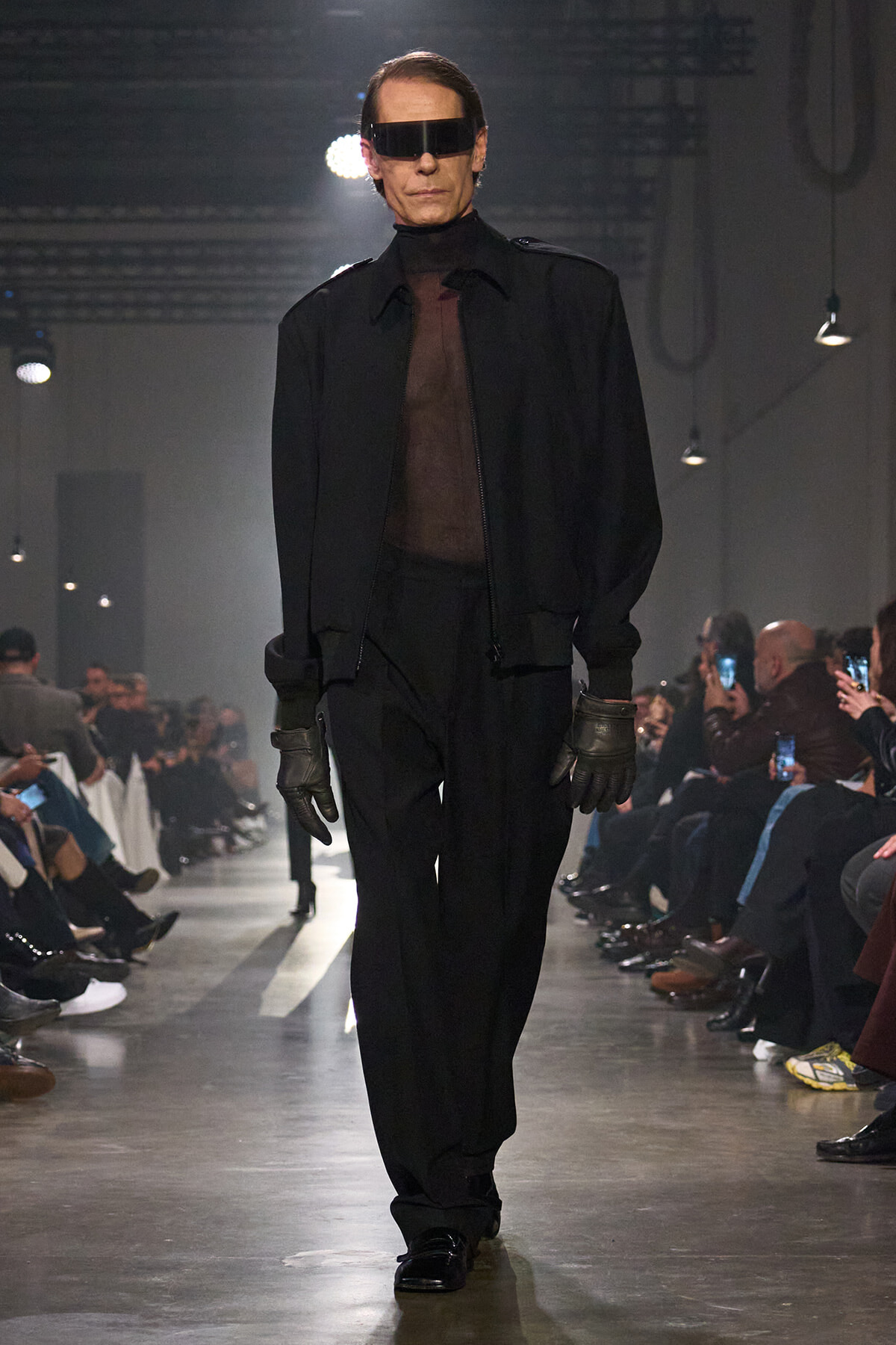 Male model on a runway in an all-black ensemble with a sheer top, zip jacket, oversized visor sunglasses, and gloves.