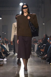 Runway model wearing a bulky brown knit sweater with a dark midi skirt, carrying a large black handbag and wearing futuristic visor sunglasses.