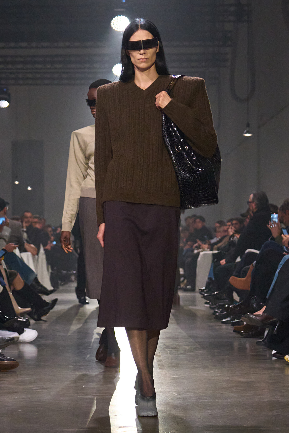 Runway model wearing a bulky brown knit sweater with a dark midi skirt, carrying a large black handbag and wearing futuristic visor sunglasses.