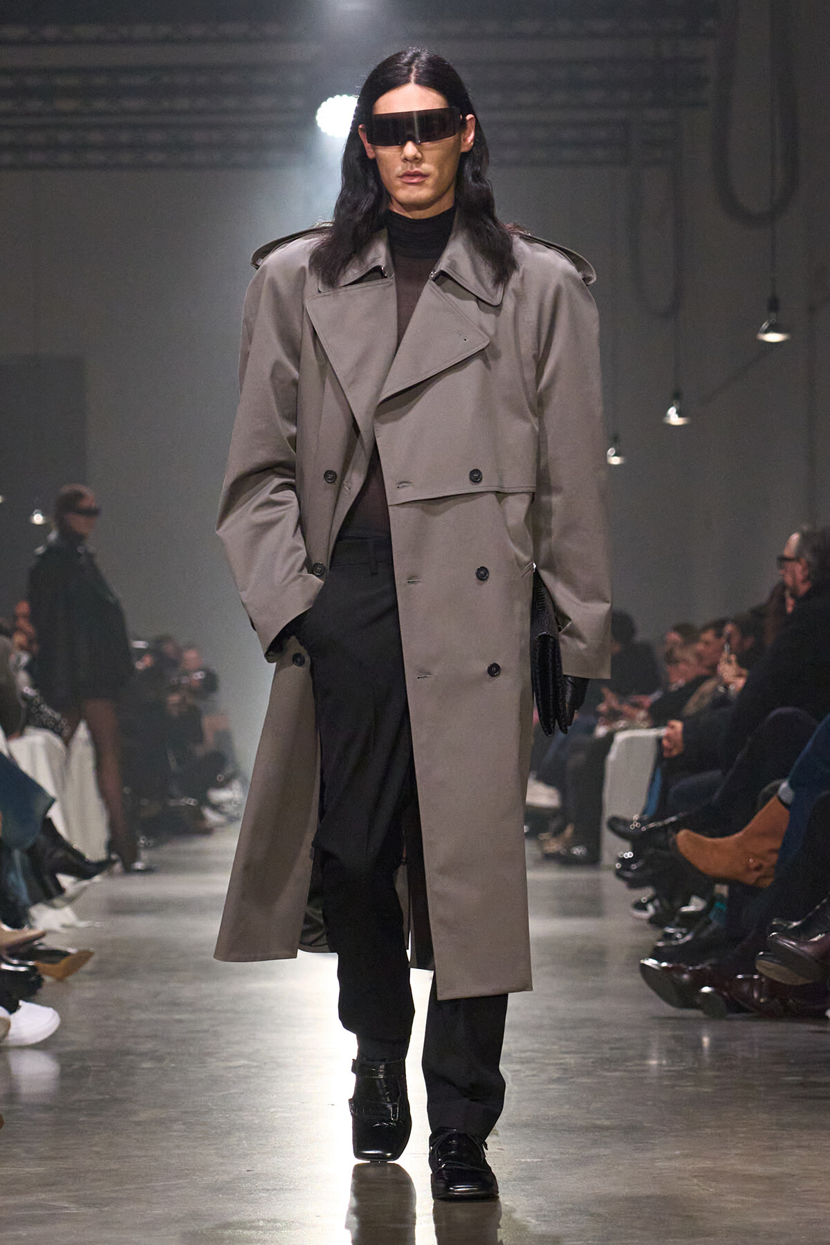 Model walks the runway in a oversized beige trench coat, black turtleneck, and futuristic rectangular sunglasses.