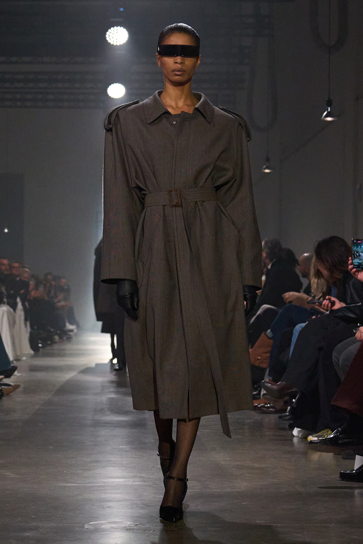 Model on a fashion runway wearing a brown belted trench coat, black gloves, and rectangular visor sunglasses; audience in background.