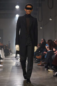 Male model walking a runway in a black tailored coat, sheer black turtleneck, black trousers, white gloves and futuristic visor sunglasses.
