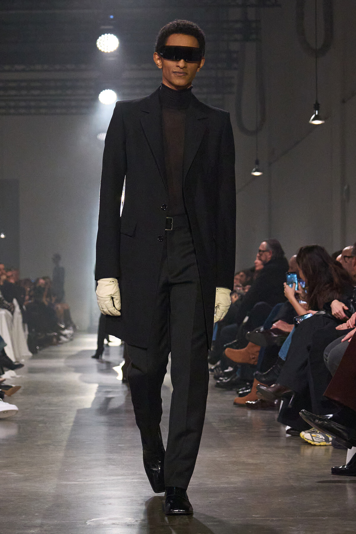 Male model walking a runway in a black tailored coat, sheer black turtleneck, black trousers, white gloves and futuristic visor sunglasses.