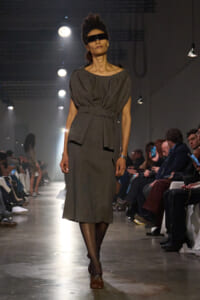 Model walks the runway in a charcoal gray belted knee-length dress with a wide neckline and black visor sunglasses, audience on both sides watching under bright lights.
