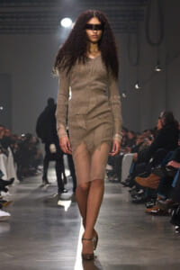 Model walks the runway in a taupe knit dress with a wide belt and layered, asymmetric skirt, wearing a black visor-style sunglasses.