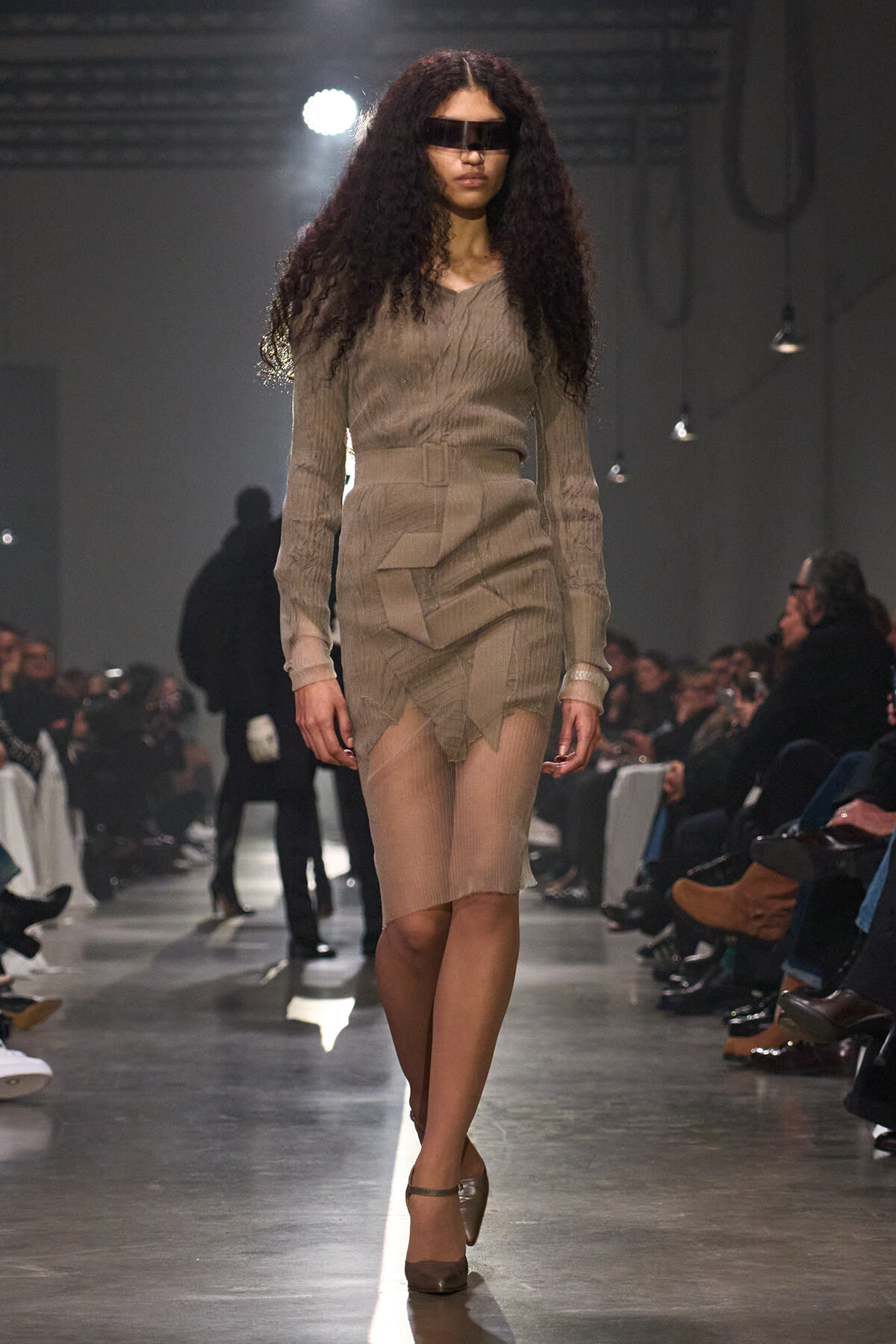 Model walks the runway in a taupe knit dress with a wide belt and layered, asymmetric skirt, wearing a black visor-style sunglasses.