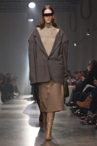 Model walking a fashion runway in a loose gray blazer over a beige turtleneck and knee-length skirt, tan boots, and futuristic eyewear.
