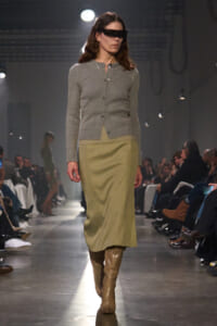 Model walks the runway in a gray cardigan, olive midi skirt, and tan knee-high boots, with futuristic black visor sunglasses.