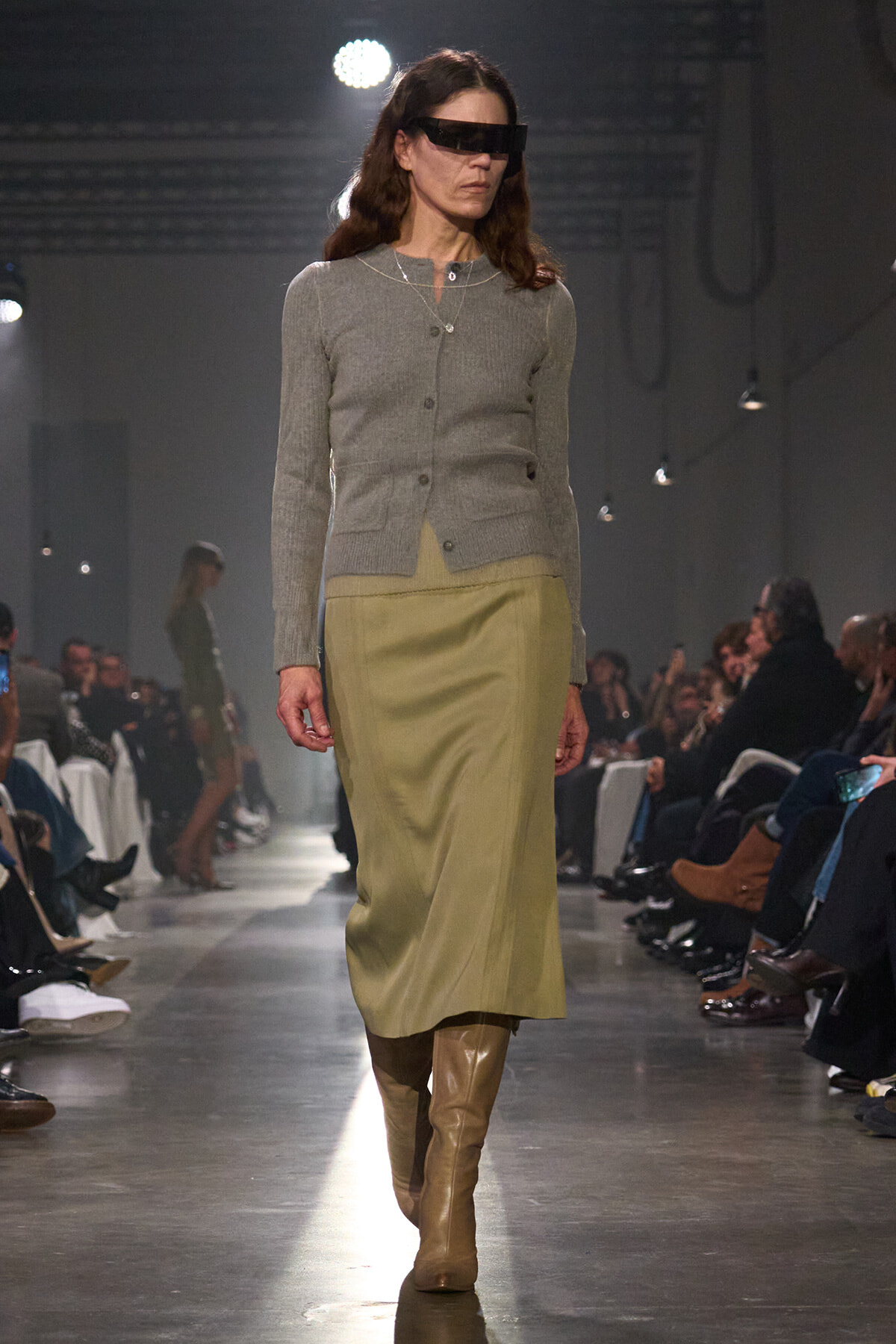 Model walks the runway in a gray cardigan, olive midi skirt, and tan knee-high boots, with futuristic black visor sunglasses.