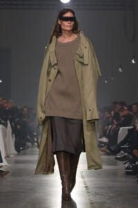 Model walks the runway in an oversized olive trench layered over a taupe knit sweater dress and brown knee-high boots, with rectangular black sunglasses.