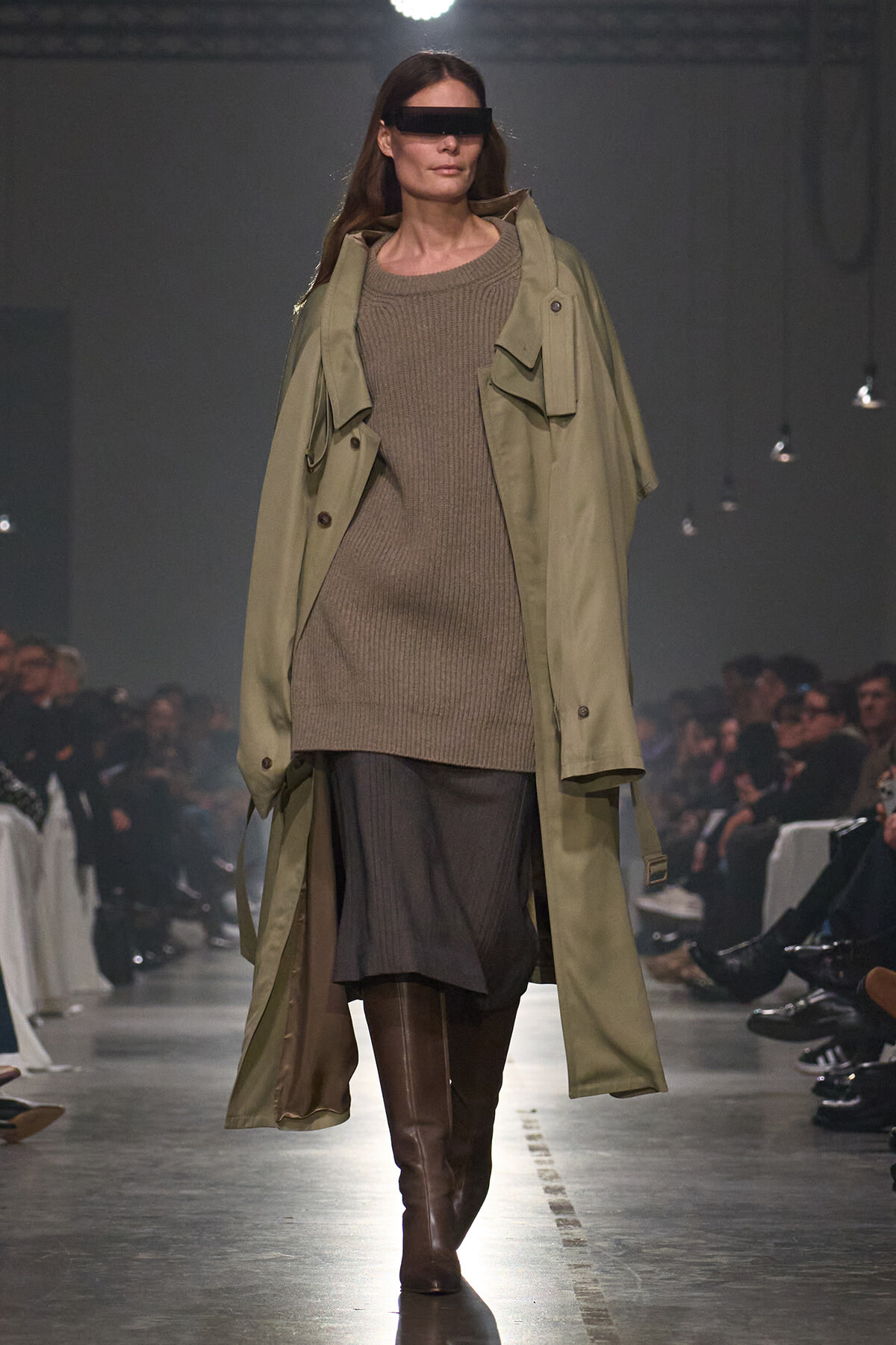 Model walks the runway in an oversized olive trench layered over a taupe knit sweater dress and brown knee-high boots, with rectangular black sunglasses.