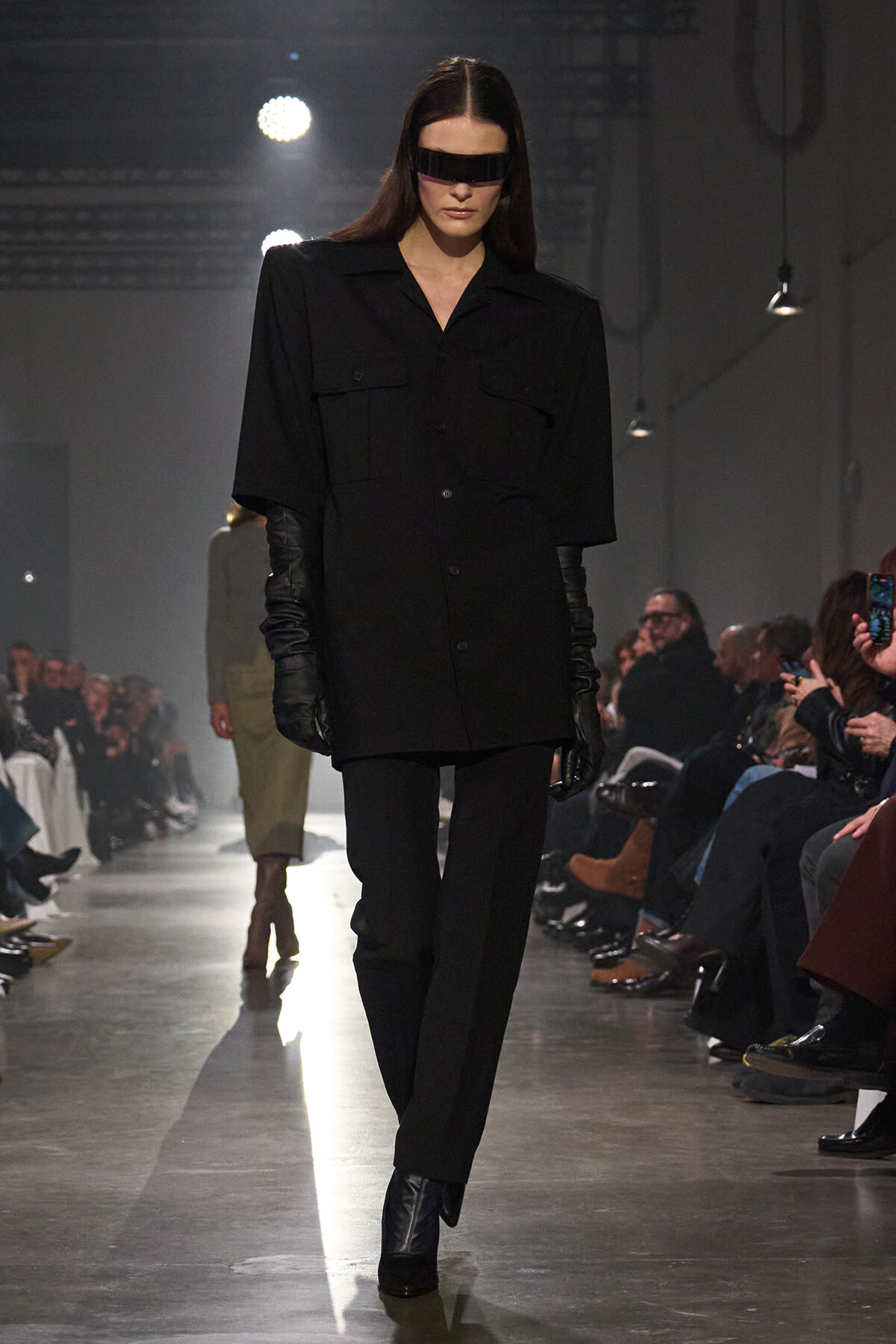 Model walks the runway in an all-black, oversized shirt-dress with long black gloves and a futuristic black visor.