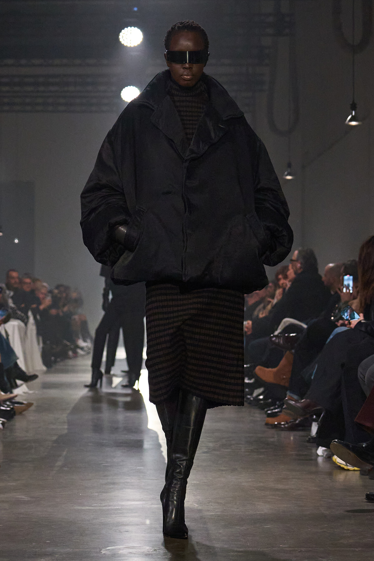 Model on runway wearing an oversized black puffer jacket, striped brown-and-black dress, tall black boots, and a futuristic dark visor.