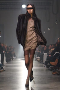 Model walks the runway in a beige buttoned dress under a dark fur coat, wearing a black blindfold and pointed black heels. Audience line the catwalk.