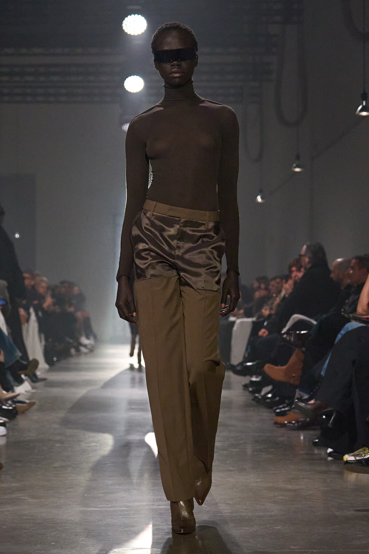 Slim model walks the runway in a turtleneck and two-tone brown pants under dramatic backstage lighting.