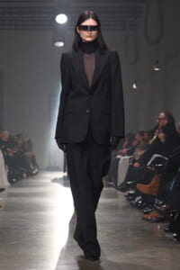 Model walks a fashion show wearing an oversized black suit with a sheer top, black gloves, and futuristic rectangular visor sunglasses on a dim runway.