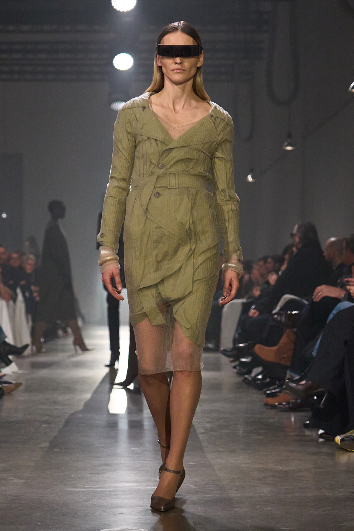 Model walks runway in olive trench dress with belt, sheer hem, and futuristic rectangular visor sunglasses.
