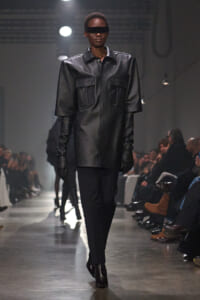 Model walking a runway in a black oversized leather coat with large pockets and gloves, wearing dark visor sunglasses.