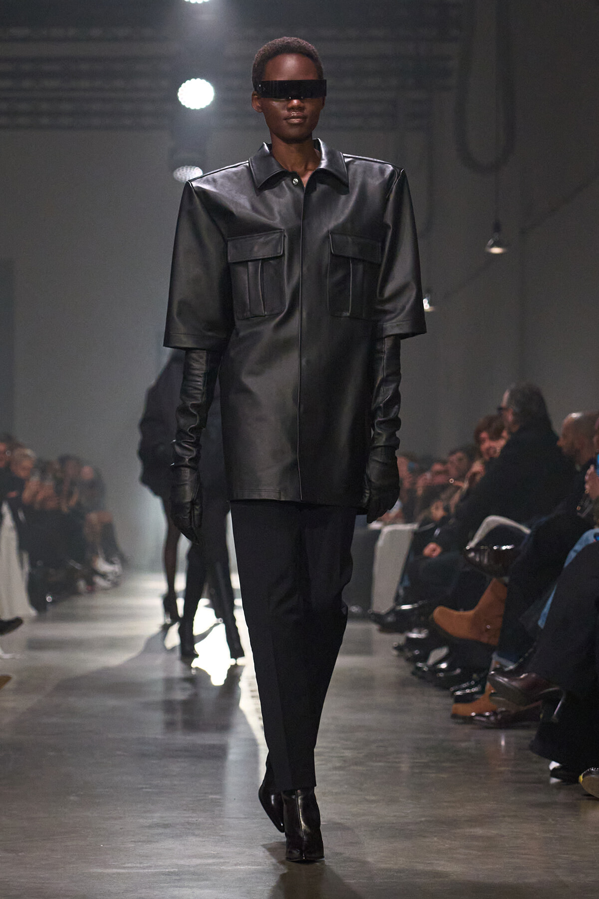 Model walking a runway in a black oversized leather coat with large pockets and gloves, wearing dark visor sunglasses.