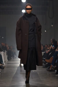 Model on a runway wearing a dark brown long coat over a black turtleneck and pleated black skirt, with black rectangular sunglasses.