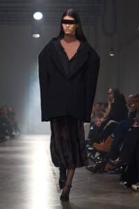 Model walking the runway in an oversized black blazer over a brown turtleneck and black fur midi skirt, wearing rectangular sunglasses that cover her eyes.