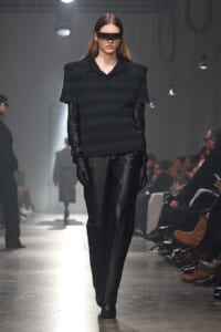 Runway model in a dark striped top, long black gloves, and futuristic visor sunglasses during a fashion show.