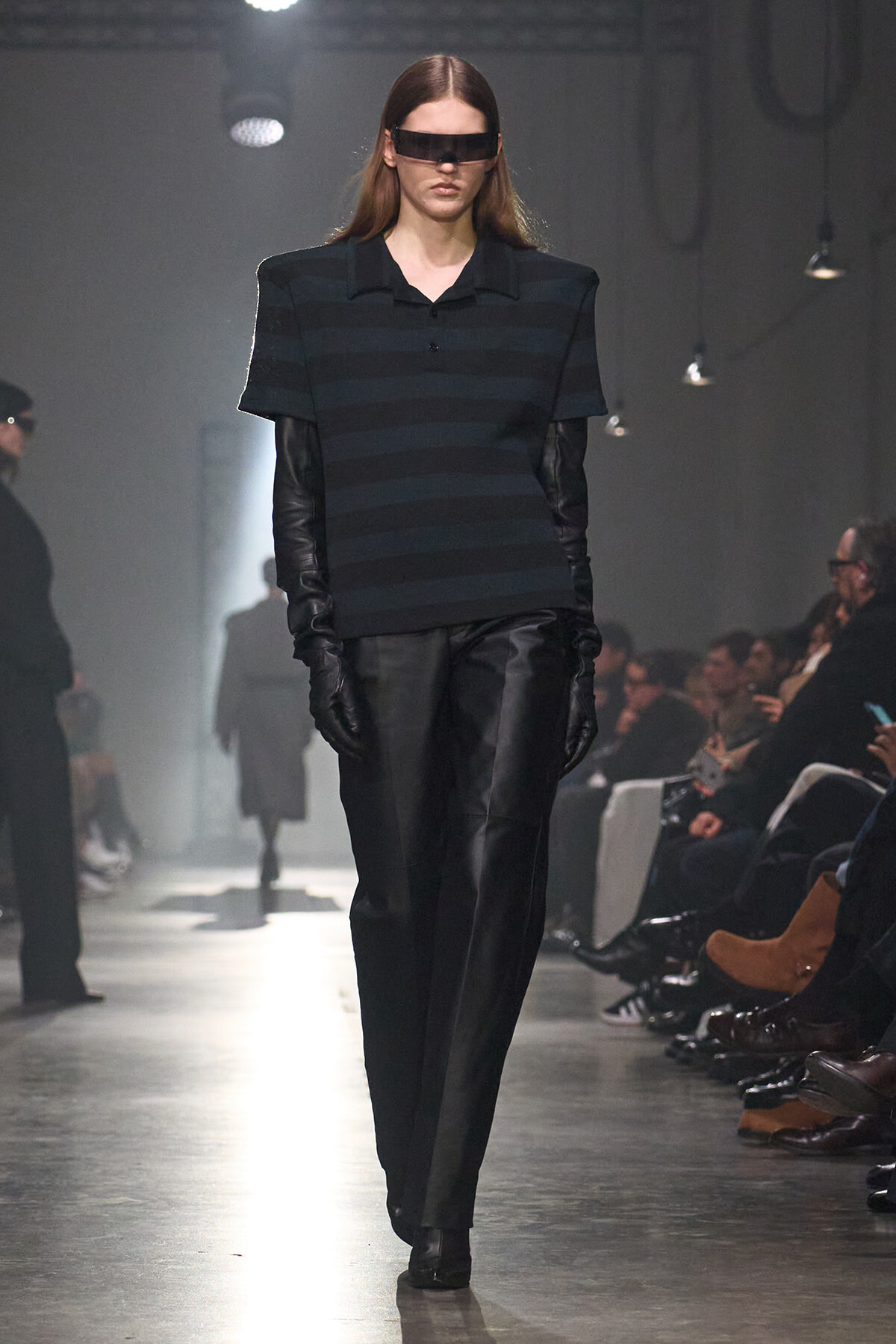 Runway model in a dark striped top, long black gloves, and futuristic visor sunglasses during a fashion show.