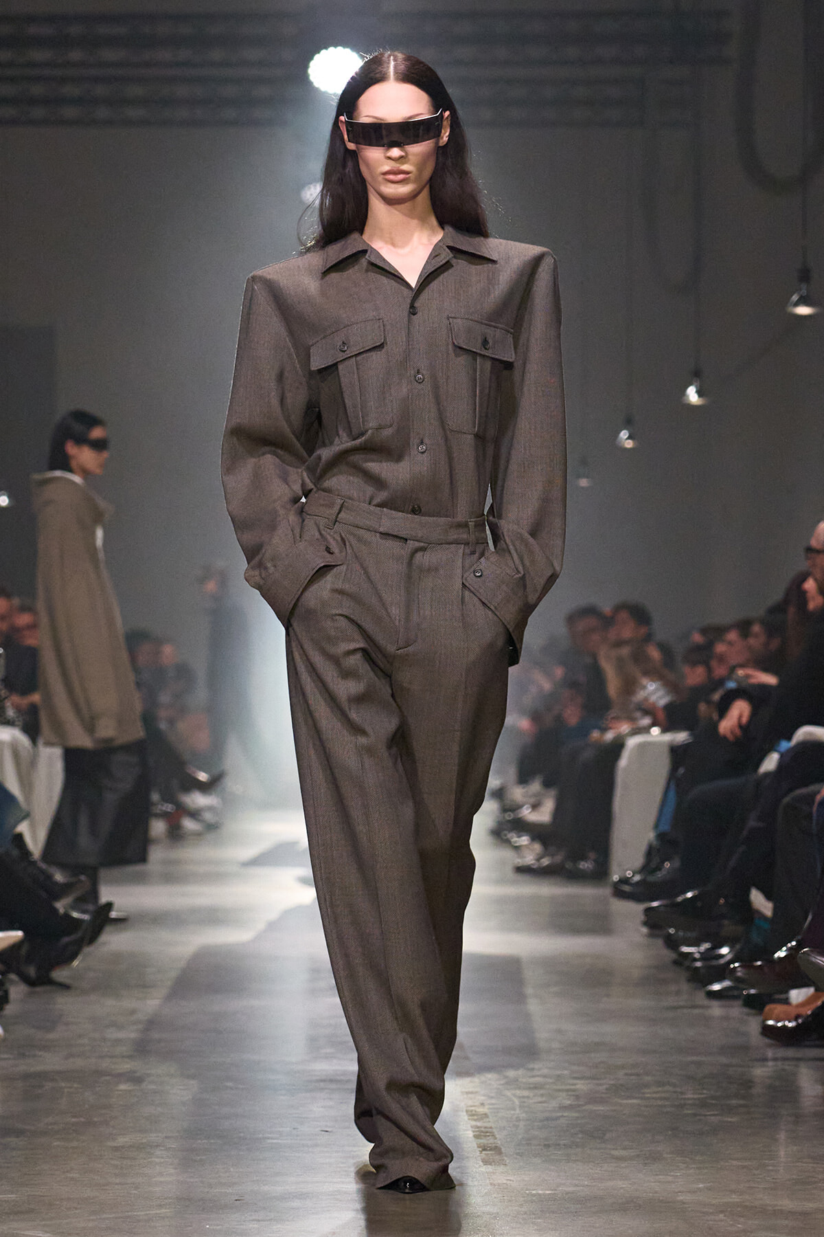 Model walking the runway in a gray pinstripe suit with oversized shoulders and a futuristic visor sunglasses.
