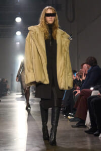 Model walking the runway in a oversized beige faux-fur coat over a black outfit with knee-high boots and a black blindfold-style headpiece.