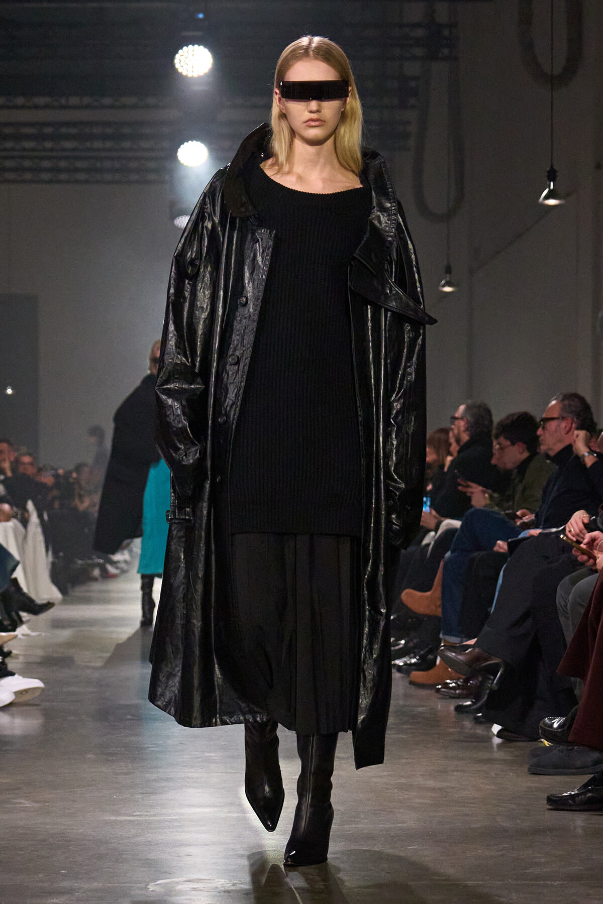 Model walks the runway in a long black patent coat over a black knit dress and knee-high boots, wearing futuristic rectangular sunglasses.