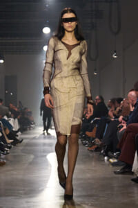 Model walks a runway in a beige pinstripe suit with sheer sleeves and brown cuffs, wearing a wide black rectangular eye-covering accessory.