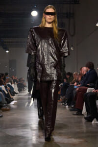 Model on a runway wearing a glossy, oversized black top and matching pants with long gloves and a futuristic rectangular visor sunglasses look.