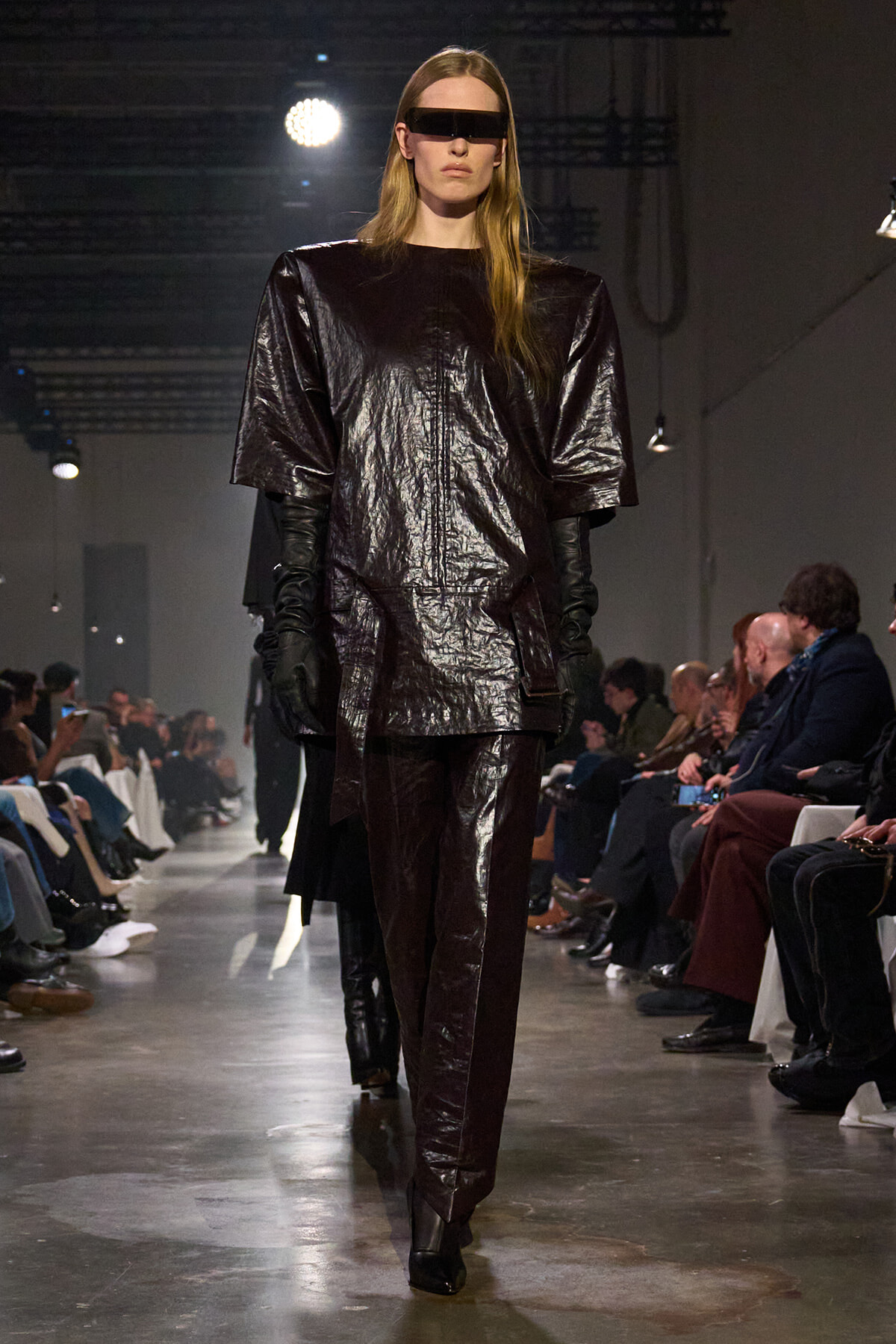 Model on a runway wearing a glossy, oversized black top and matching pants with long gloves and a futuristic rectangular visor sunglasses look.