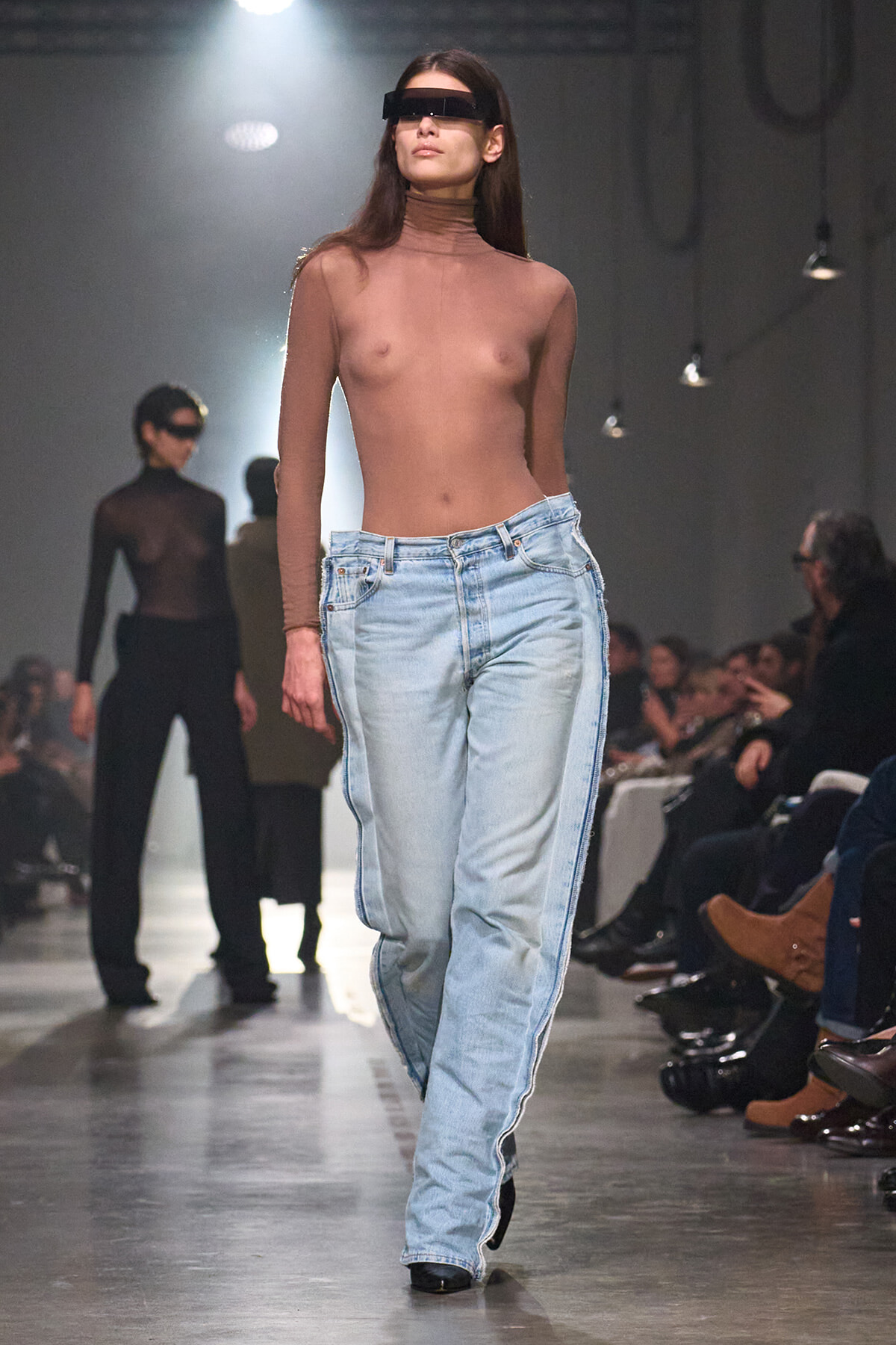 Model walking a runway in a sheer brown long-sleeve top, light-wash jeans, and a futuristic black visor-style sunglasses.