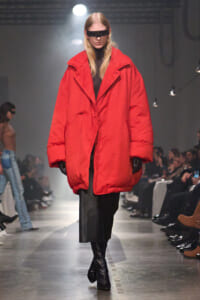 Model walking a runway in a bright red oversized puffer coat, black gloves, and futuristic rectangular sunglasses.
