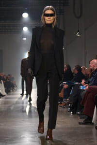 Model walking a dim runway in an all-black outfit: sheer top, oversized blazer, slim pants, brown ankle boots, black gloves, and a futuristic rectangular visor.